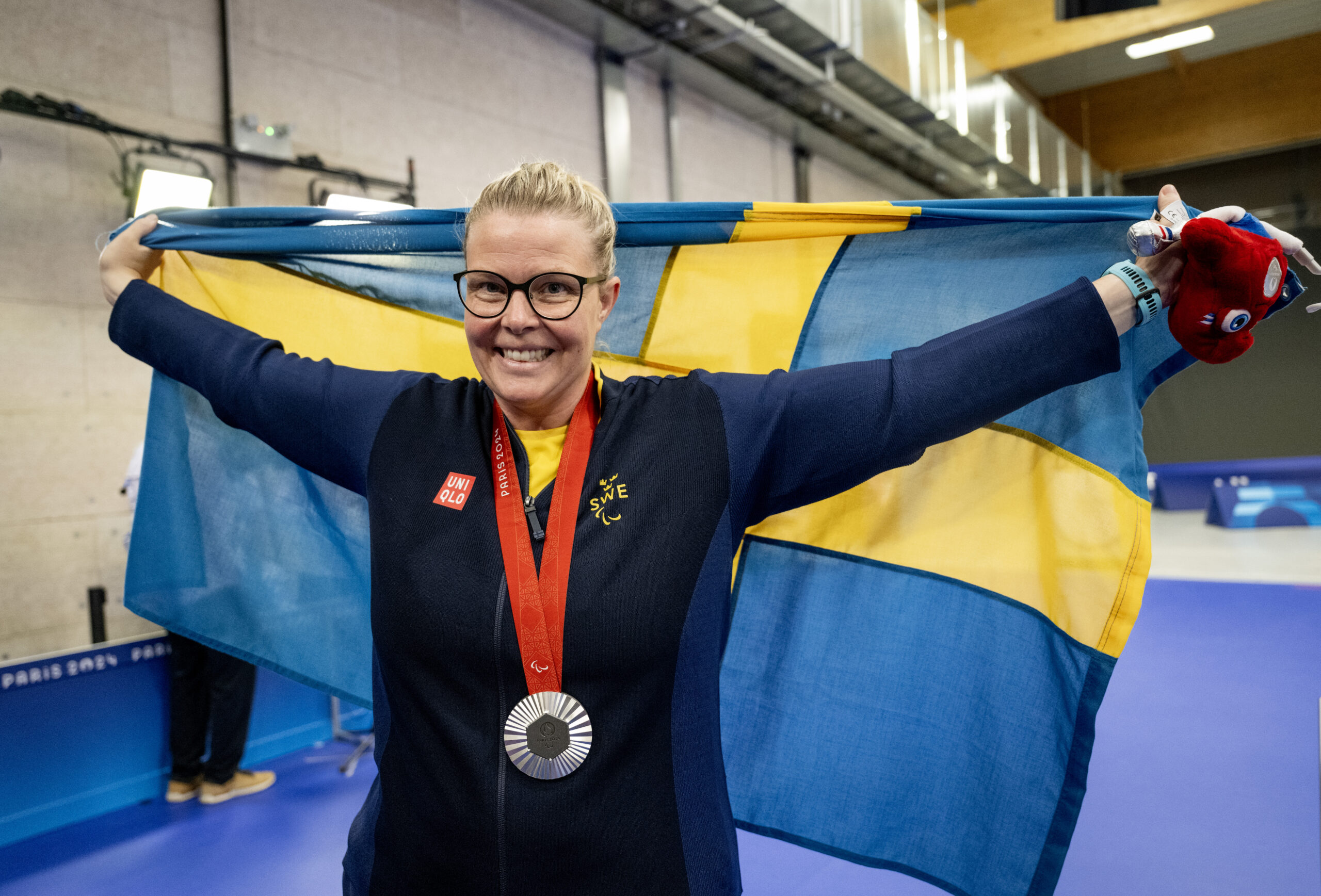 Anna Benson in a gym with her silver medal on, holding the Swedish flag and a lucky mascot.