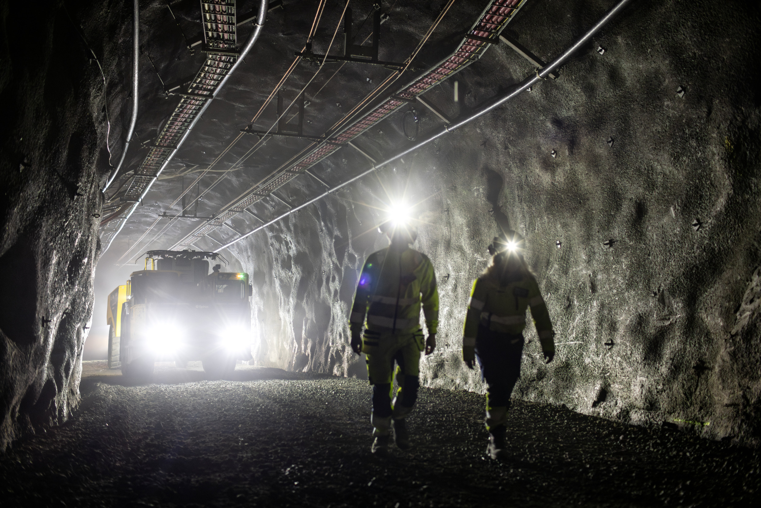 Two workers walk in front of a heavy-duty electric vehicle in an underground mine tunnel.